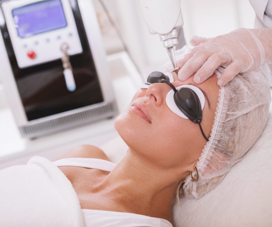 Close up of a beautiful woman in protective eyeglasses getting facial laser treatment by professional cosmetologist